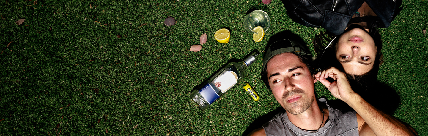 Guy and Girl laying in grass listening to music, crystal breth and bottle of alcohol laying in grass next to them. 