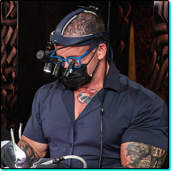 Dentist performing procedure glasses tattoos head light sitting next to patient